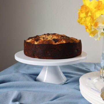 A rustic French apple and chocolate cake on a white pedestal cake stand, set against a soft blue linen and a bouquet of yellow daffodils in the background. The cake has a golden, textured top with visible apple slices and melted chocolate pieces.