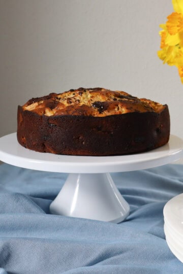 A rustic French apple and chocolate cake on a white pedestal cake stand, set against a soft blue linen and a bouquet of yellow daffodils in the background. The cake has a golden, textured top with visible apple slices and melted chocolate pieces.