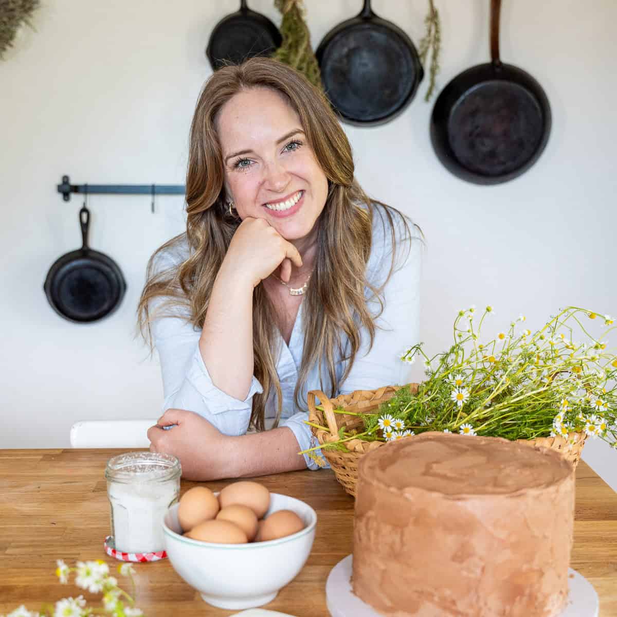 Woman in a rustic kitchen with chocolate cake, eggs, and chamomile flowers on table.