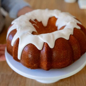 Vanilla Glaze perfectly drips down the sides of a vanilla Bundt Cake on a white cake stand sitting on a butcher block table