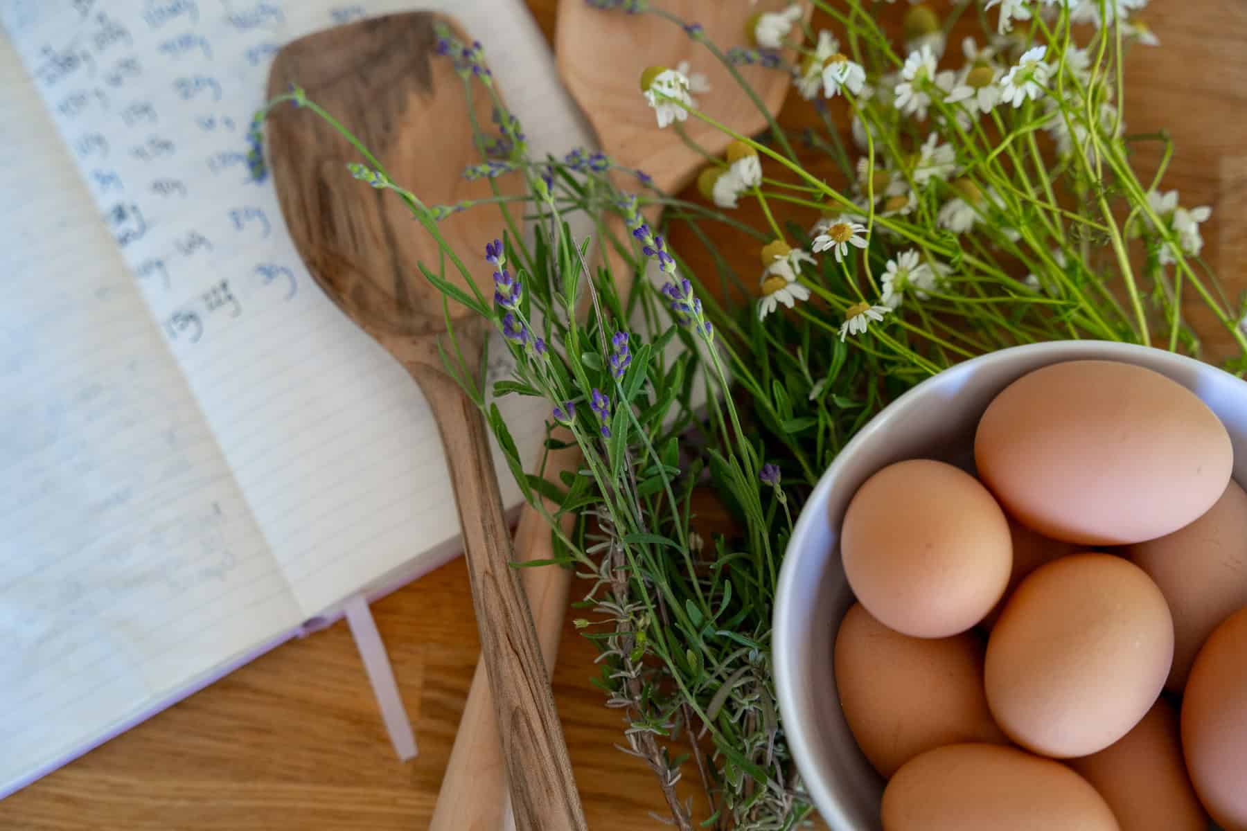 bowl of brown eggs in a French porcelain bowl with fresh flowers and lavender with a recipe notebook and handmade wooden spoons