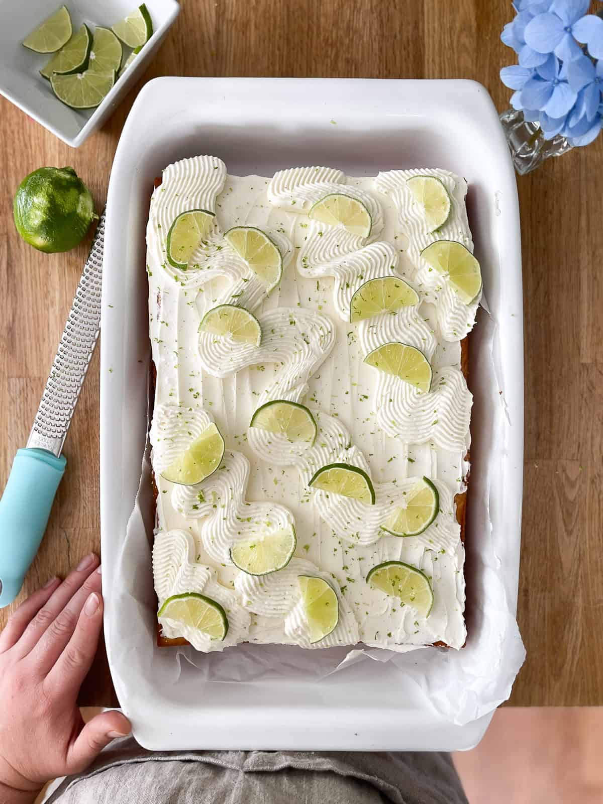 A golden vanilla sheet cake topped with swirls of lime-kissed frosting, garnished with fresh lime slices and a dusting of zest, set in a white baking dish on a wooden table with a lime and zester nearby.