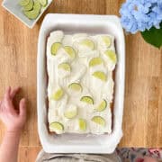 A golden vanilla sheet cake topped with swirls of lime-kissed frosting, garnished with fresh lime slices and a dusting of zest, set in a white baking dish on a wooden table with a lime and zester nearby.