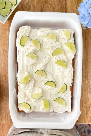 A golden vanilla sheet cake topped with swirls of lime-kissed frosting, garnished with fresh lime slices and a dusting of zest, set in a white baking dish on a wooden table with a lime and zester nearby.