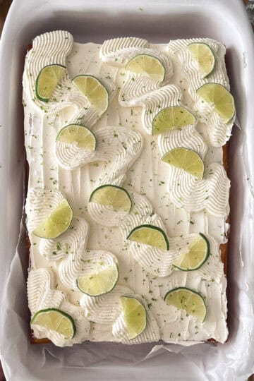 A golden vanilla sheet cake topped with swirls of lime-kissed frosting, garnished with fresh lime slices and a dusting of zest, set in a white baking dish on a wooden table with a lime and zester nearby.