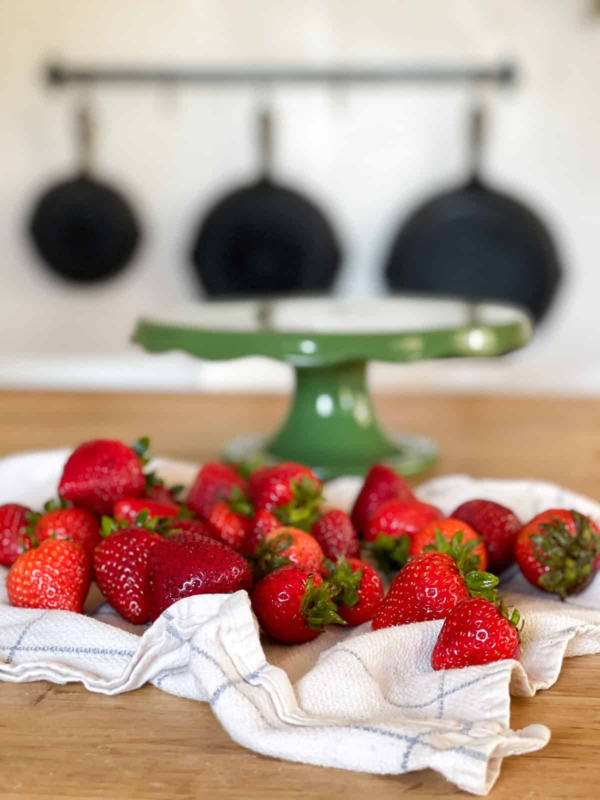 fresh strawberries on a blue striped towel with a vintage green cake stand in the background on a wooden counter, behind that are cast iron skillets hanging on the wall