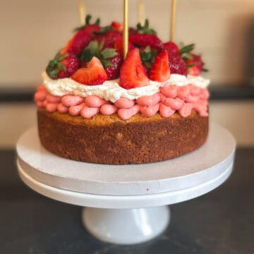 A rustic single layer shortcake cake layered with strawberry buttercream frsoting and whipped cream topped with fresh strawberries on a white cake place on the a black counter top.