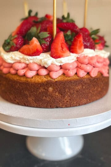 A rustic single layer shortcake cake layered with strawberry buttercream frsoting and whipped cream topped with fresh strawberries on a white cake place on the a black counter top.