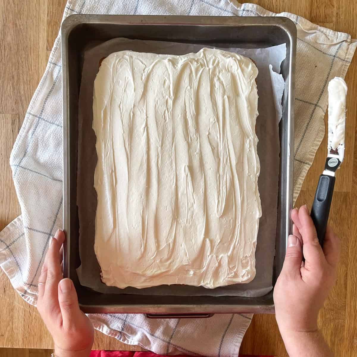 Hands holding a metal pan with a 9x13 rectangle vanilla sheet cake topped with smooth homemade vanilla frosting. The cake rests on a towel-covered wooden surface, with an offset spatula beside it.