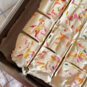 Close-up of a 9x13 vanilla sheet cake sliced into squares and topped with creamy white vanilla frosting and colorful rainbow sprinkles, sitting in a metal baking pan lined with parchment paper.