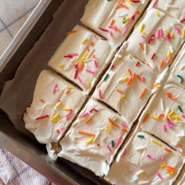 Close-up of a 9x13 vanilla sheet cake sliced into squares and topped with creamy white vanilla frosting and colorful rainbow sprinkles, sitting in a metal baking pan lined with parchment paper.