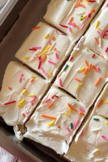 Close-up of a 9x13 vanilla sheet cake sliced into squares and topped with creamy white vanilla frosting and colorful rainbow sprinkles, sitting in a metal baking pan lined with parchment paper.