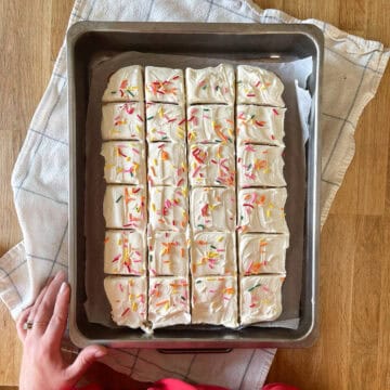 Vanilla frosted sheet cake in a rustic cake pan over a towel with baker's hands nearby