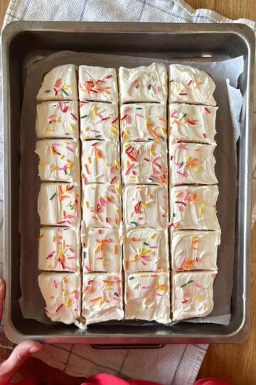Vanilla frosted sheet cake in a rustic cake pan over a towel with baker's hands nearby