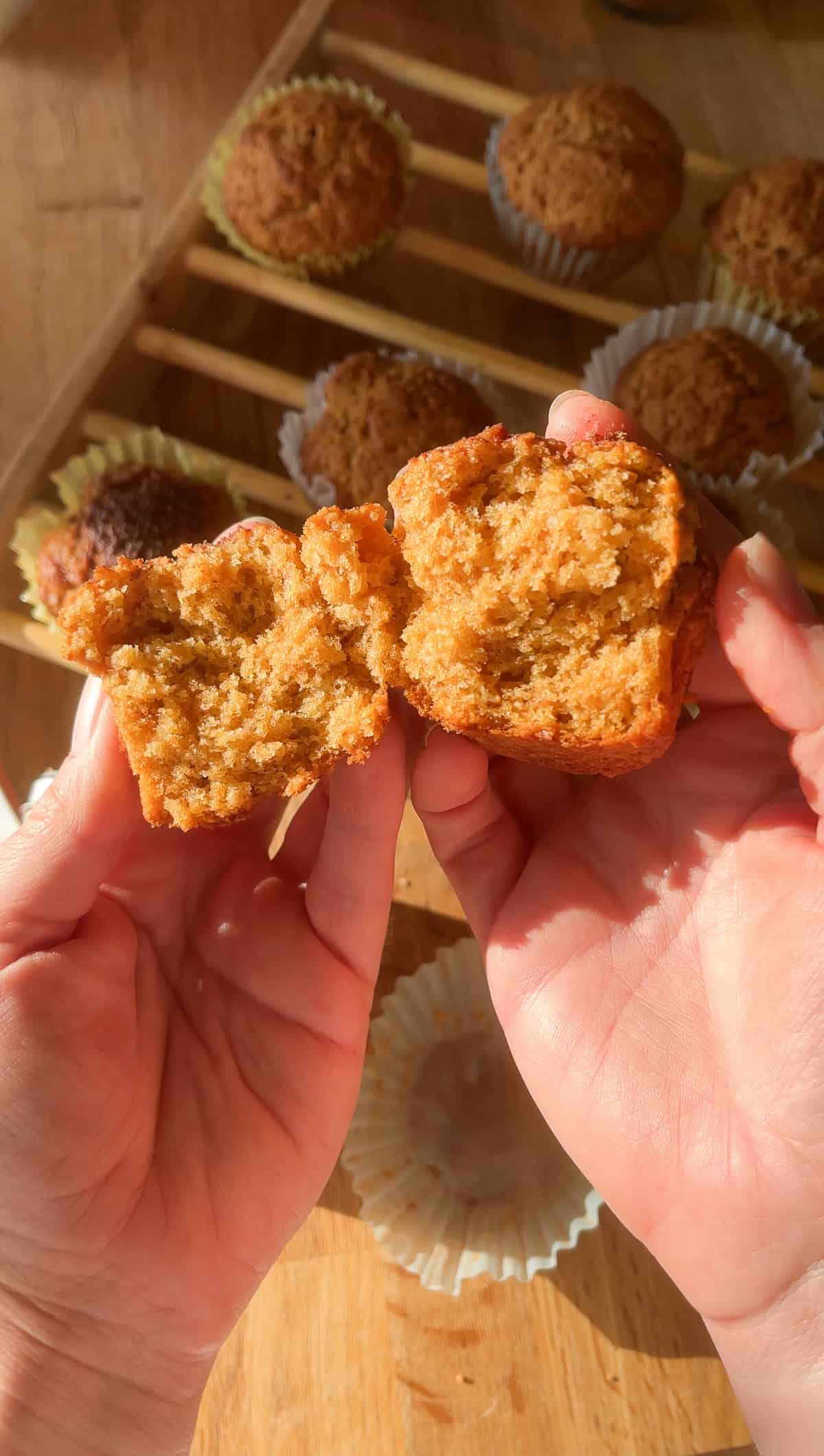 Split open Golden brown French breakfast muffins cooling on a wooden rack, with soft sunlight highlighting their rustic, cinnamon-sugar tops and crinkled white paper liners.
