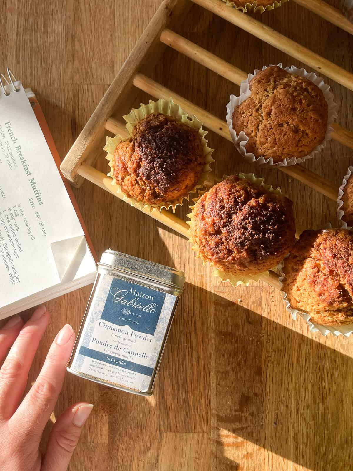 Golden brown French breakfast muffins cooling on a wooden rack, with soft sunlight highlighting their rustic, cinnamon-sugar tops and crinkled white paper liners with tin of cinnamon nearby