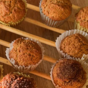 Golden brown French breakfast muffins cooling on a wooden rack, with soft sunlight highlighting their rustic, cinnamon-sugar tops and crinkled white paper liners.