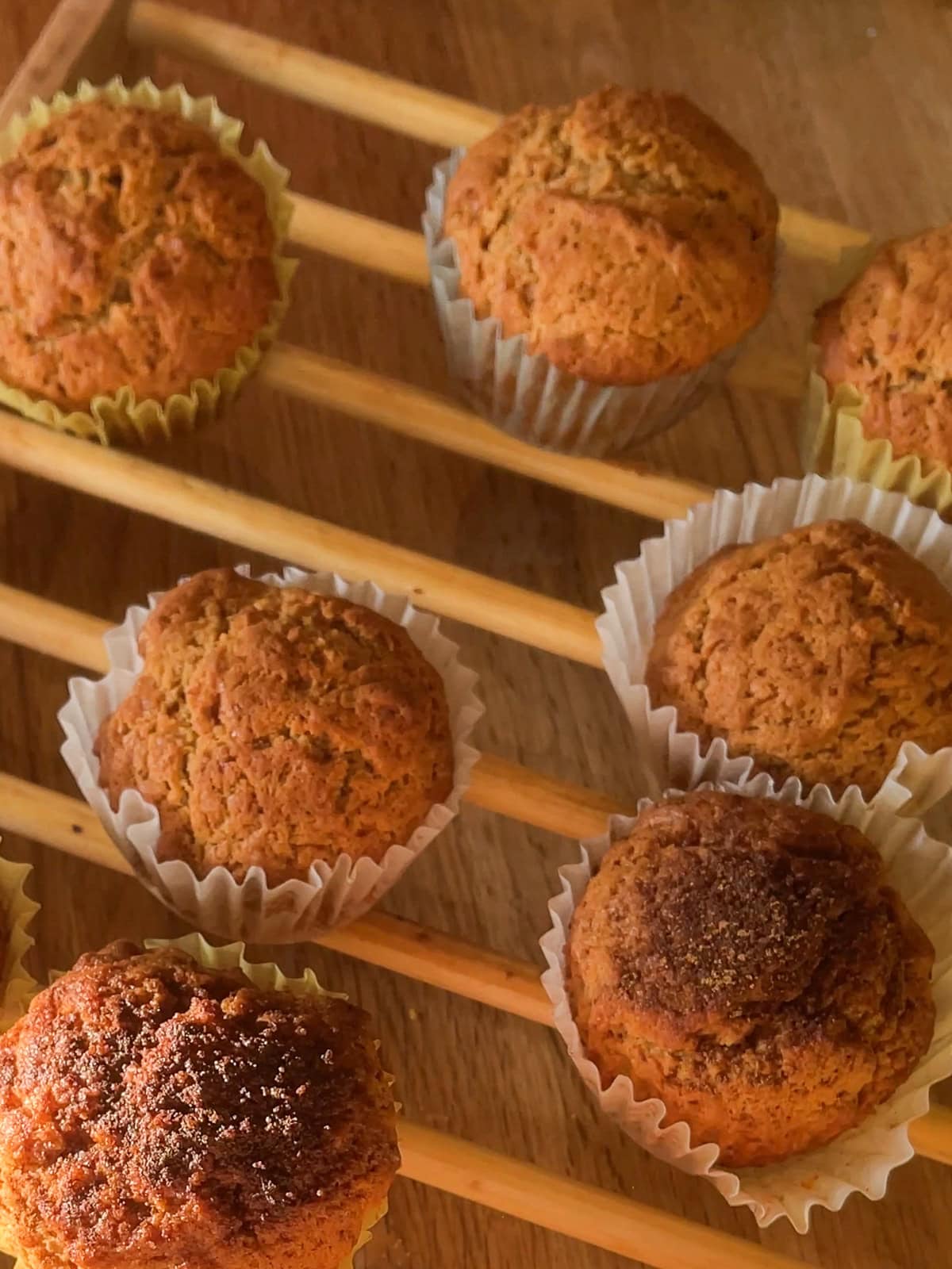 Golden brown French breakfast muffins cooling on a wooden rack, with soft sunlight highlighting their rustic, cinnamon-sugar tops and crinkled white paper liners.