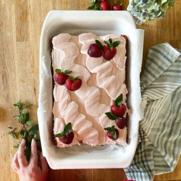 A vanilla sheet cake topped with piped swirls of strawberry frosting and garnished with fresh strawberries and mint leaves, served in a white baking dish on a wood surface with a striped kitchen towel and greenery nearby.