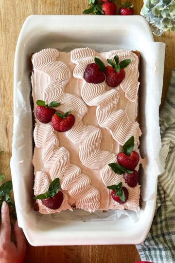 A vanilla sheet cake topped with piped swirls of strawberry frosting and garnished with fresh strawberries and mint leaves, served in a white baking dish on a wood surface with a striped kitchen towel and greenery nearby.
