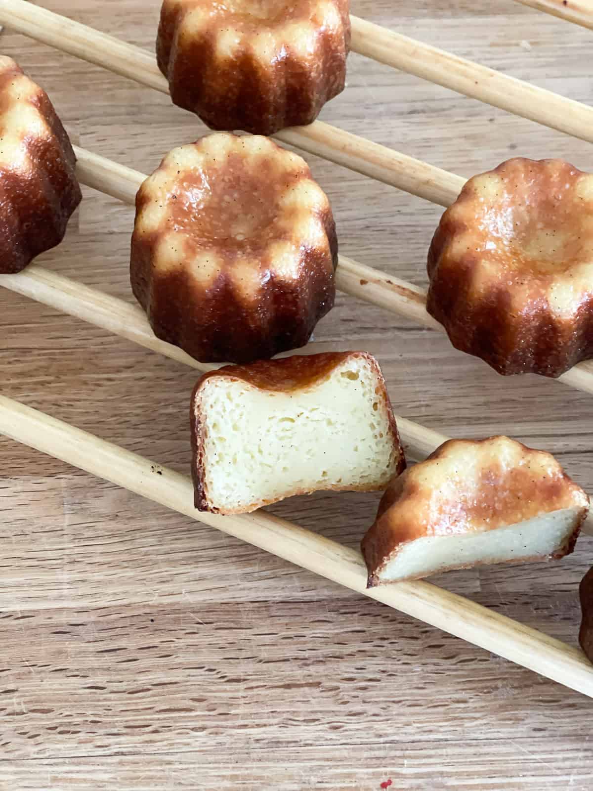 Freshly baked canelés with glossy, caramelized crusts cooling on a wooden rack, with one sliced open to reveal its tender, custardy vanilla interior.