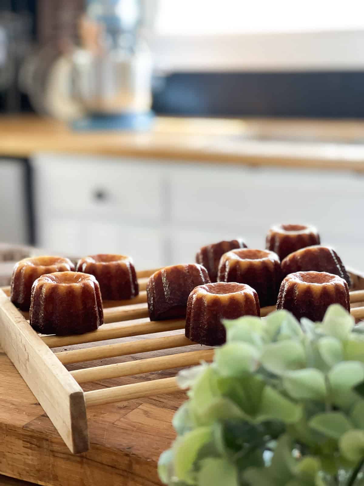 Freshly baked canelés de Bordeaux with caramelized crusts cooling on a wooden rack in a bright countryside kitchen, with a blurred hydrangea in the foreground.