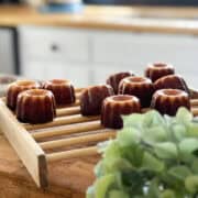 Freshly baked canelés de Bordeaux with caramelized crusts cooling on a wooden rack in a bright countryside kitchen, with a blurred hydrangea in the foreground.
