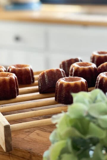 Freshly baked canelés de Bordeaux with caramelized crusts cooling on a wooden rack in a bright countryside kitchen, with a blurred hydrangea in the foreground.