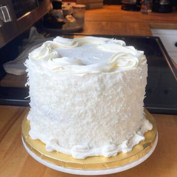 A close-up of a finished three-layer coconut cake on a kitchen counter, coated in shredded coconut with piped buttercream borders and a smooth frosted top.