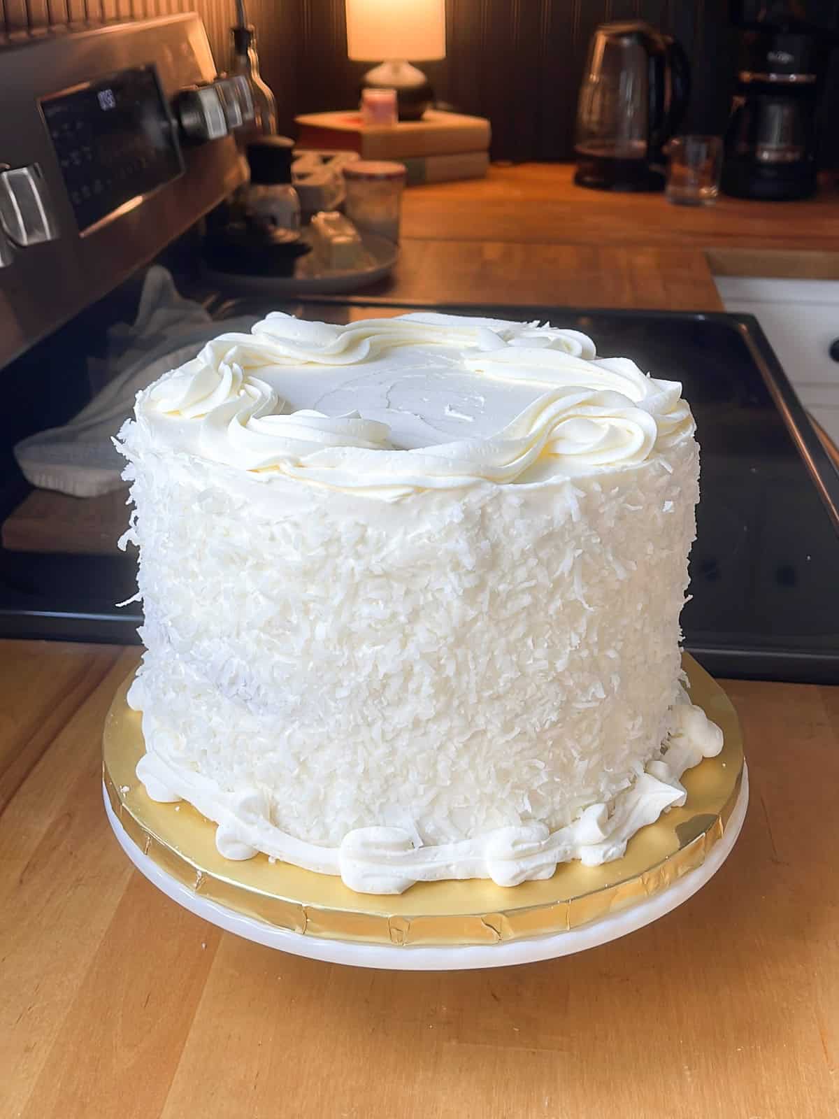 A close-up of a finished three-layer coconut cake on a kitchen counter, coated in shredded coconut with piped buttercream borders and a smooth frosted top.