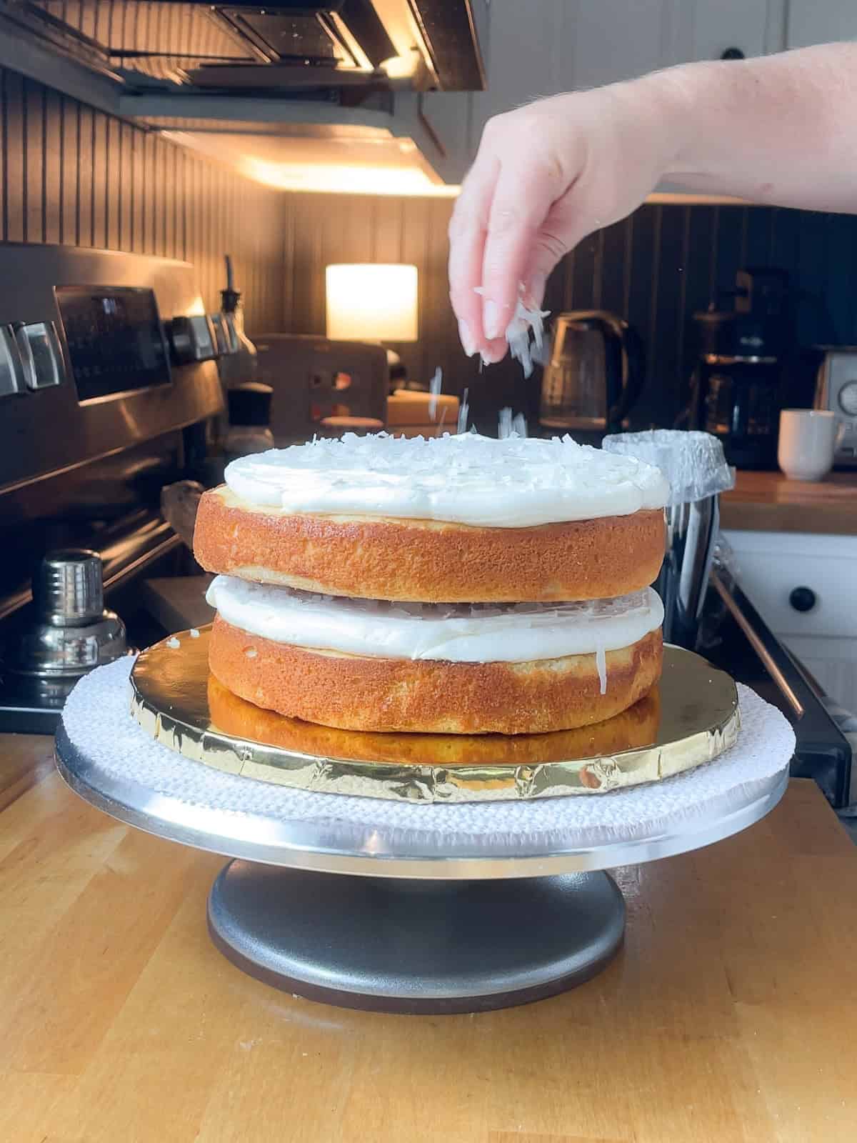 A hand sprinkles shredded coconut over the buttercream layer of a stacked, unfrosted three-layer cake on a rotating cake stand in a cozy kitchen setting.
