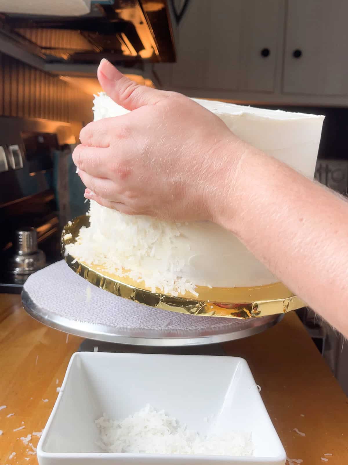 A hand applies shredded coconut to the sides of a buttercream 3 layer cake on a rotating cake stand in a cozy kitchen setting.