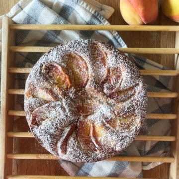sugar dusted rustic peach cobbler cake on a wooden cooling rack with a blue striped towel and fresh peaches on a wooden surface