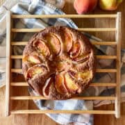 sugar dusted rustic peach cobbler cake on a wooden cooling rack with a blue striped towel and fresh peaches on a wooden surface