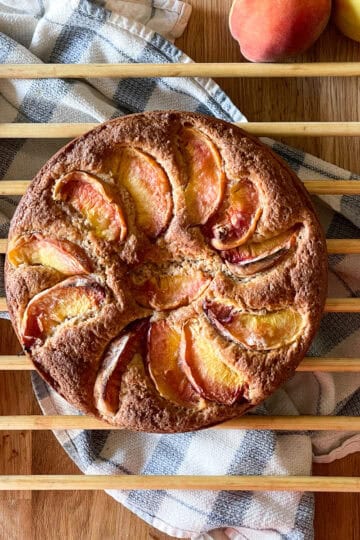 sugar dusted rustic peach cobbler cake on a wooden cooling rack with a blue striped towel and fresh peaches on a wooden surface