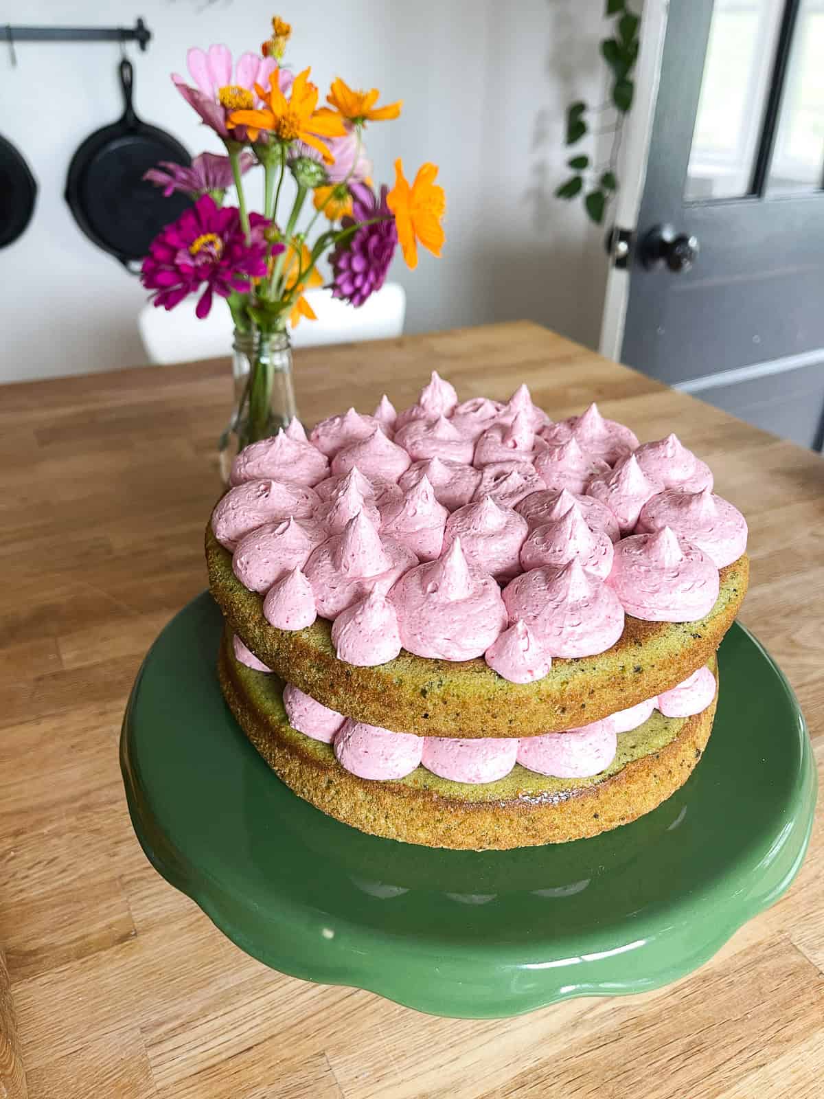 A rustic two-layer matcha cake is filled and topped with pink raspberry frosting. The cake is displayed on a glossy green pedestal cake stand, with a wooden table and blurred flowers in the background.