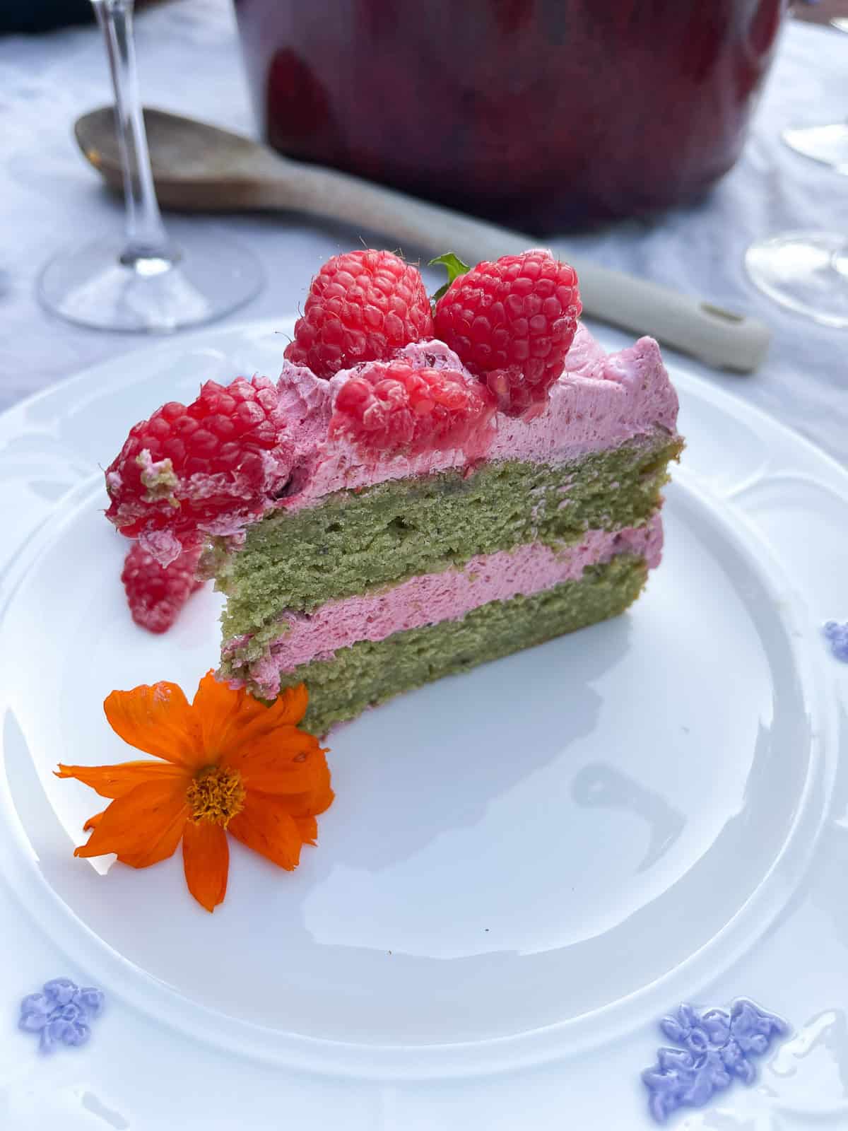 A slice of layer cake with bright green matcha cake layers and a bright pink raspberry frosting with a fresh cosmo flower bud, topped with fresh raspberries on a white china plate