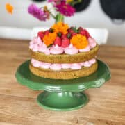 A rustic two-layer matcha cake is filled and topped with pink raspberry frosting, fresh raspberries, and vibrant orange edible flowers. The cake is displayed on a glossy green pedestal cake stand, with a wooden table and blurred flowers in the background.