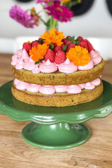 A rustic two-layer matcha cake is filled and topped with pink raspberry frosting, fresh raspberries, and vibrant orange edible flowers. The cake is displayed on a glossy green pedestal cake stand, with a wooden table and blurred flowers in the background.