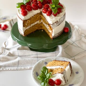 A two layer rustic tomato cake with white basil frosting naked-style with fresh cherry tomatoes and fresh basil bouquets sitting on top. The cake sits on a vintage green cake stand, with a slice of cake sitting on white china in front.