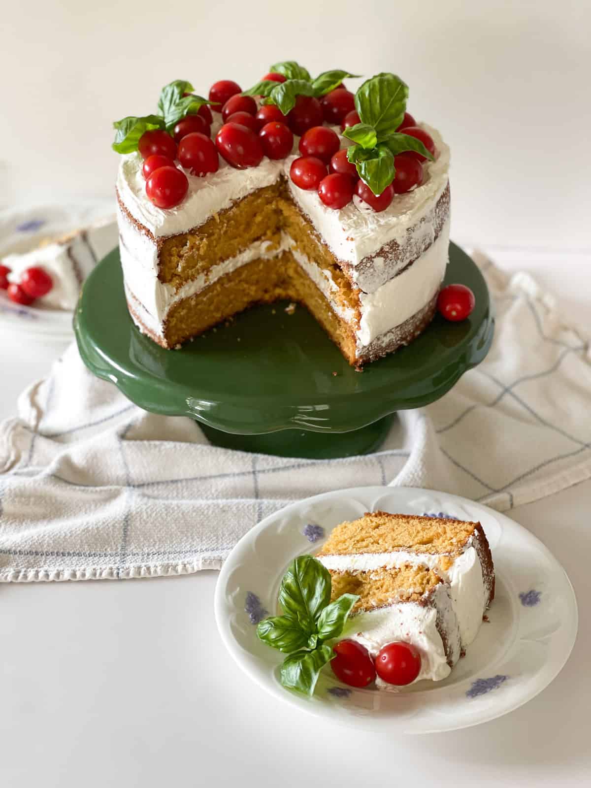 A two layer rustic tomato cake with white basil frosting naked-style with fresh cherry tomatoes and fresh basil bouquets sitting on top. The cake sits on a vintage green cake stand, with a slice of cake sitting on white china in front.