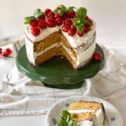 A two layer rustic tomato cake with white basil frosting naked-style with fresh cherry tomatoes and fresh basil bouquets sitting on top. The cake sits on a vintage green cake stand, with a slice of cake sitting on white china in front.