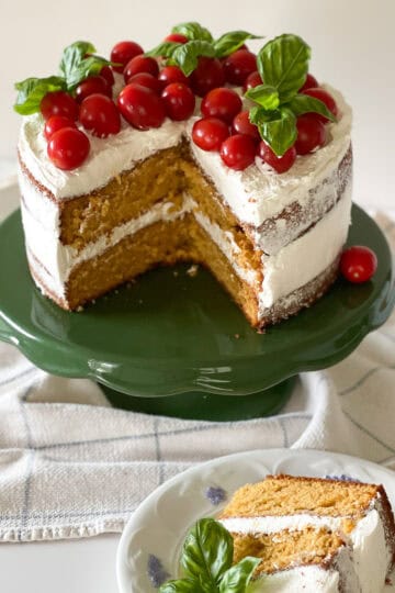 A two layer rustic tomato cake with white basil frosting naked-style with fresh cherry tomatoes and fresh basil bouquets sitting on top. The cake sits on a vintage green cake stand, with a slice of cake sitting on white china in front.