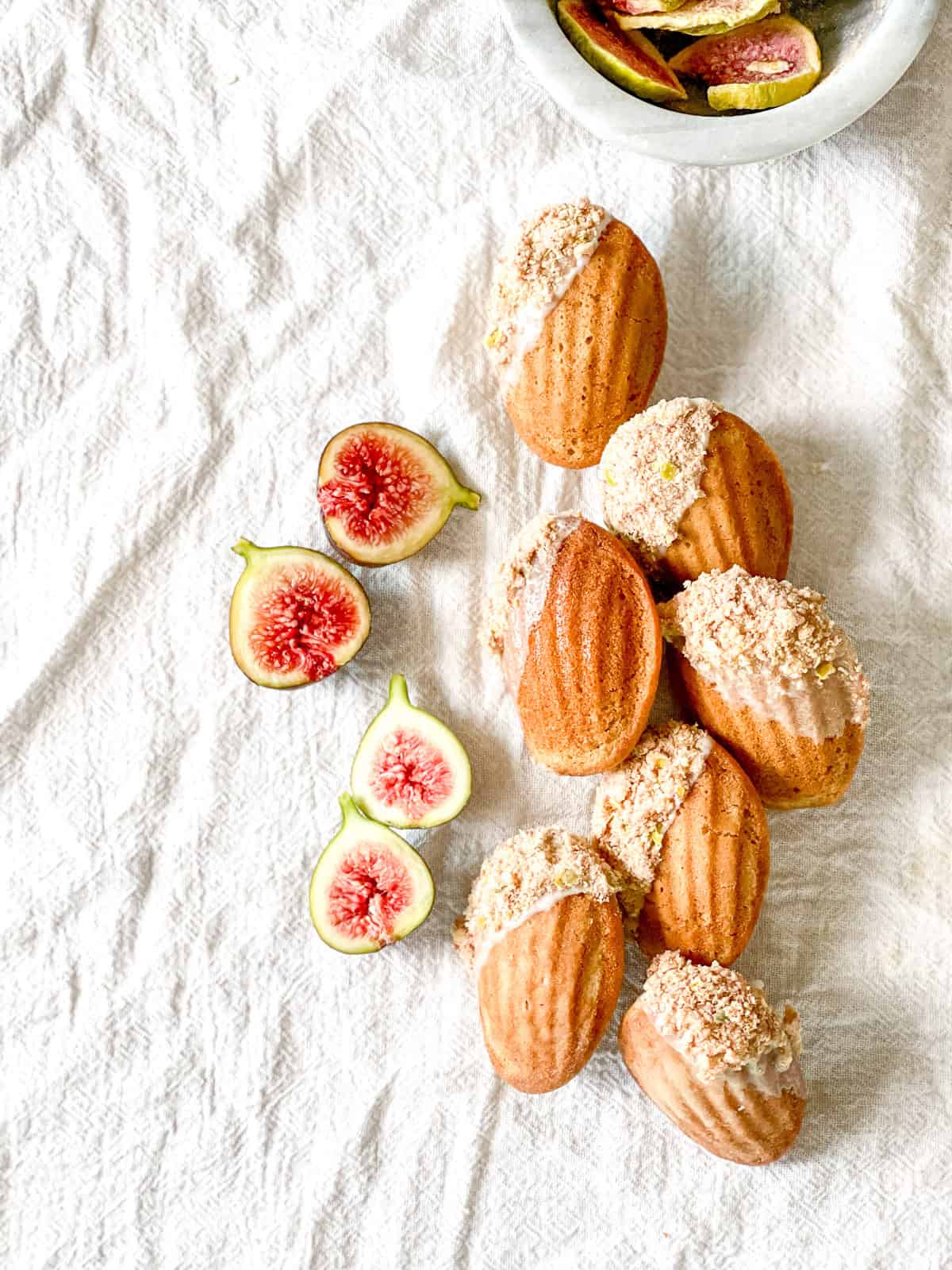 Golden brown spiced madeleines with a soft shell pattern and a partial dip into a white glaze and then a coating of freeze-dried figs all arranged on a white linen cloth.
