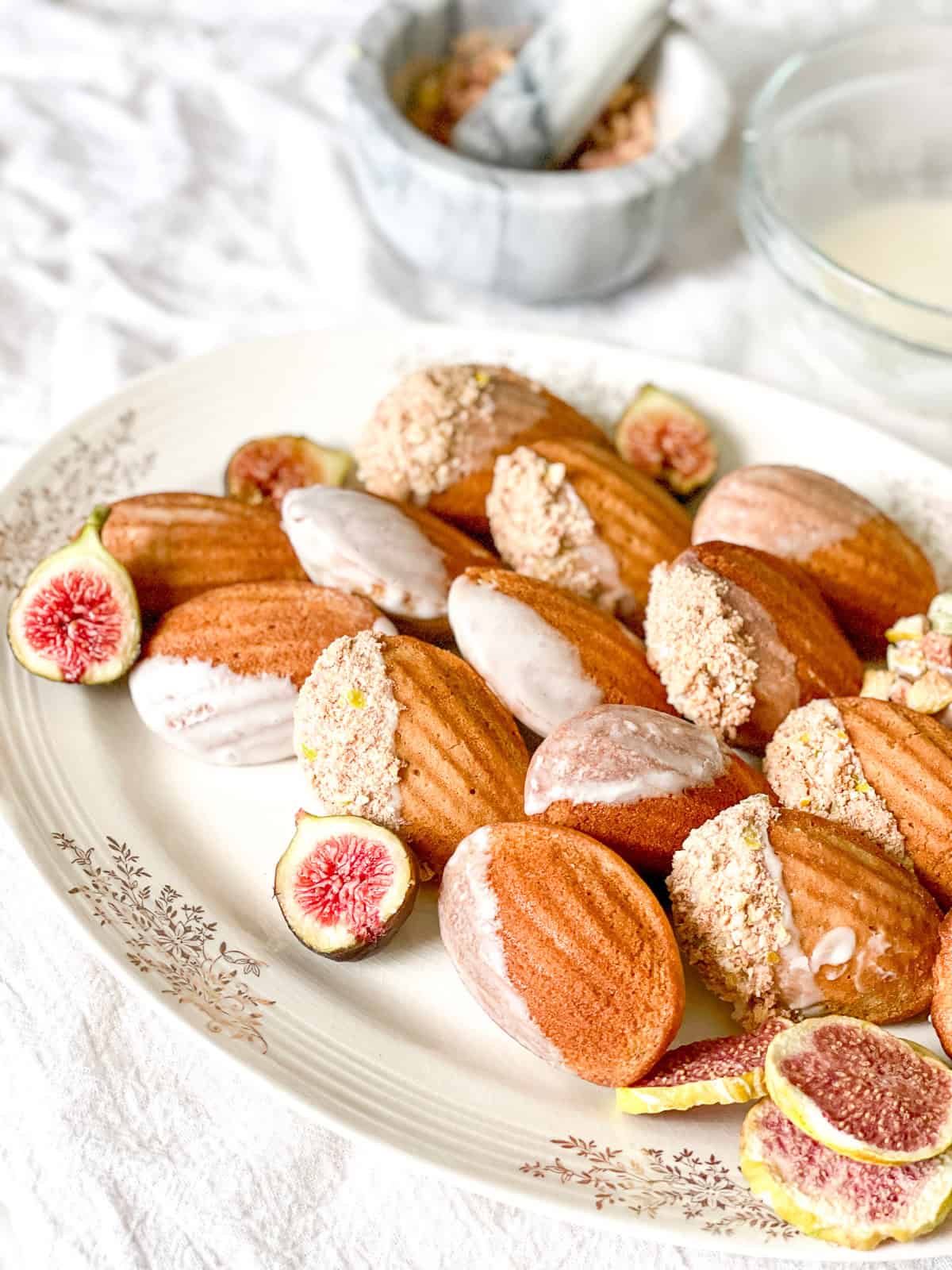 Golden brown spiced madeleines with a soft shell pattern and a partial dip into a white glaze and then a coating of freeze-dried figs all arranged on an elegant porcelain plate sitting on white linen cloth.
