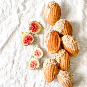 Golden brown spiced madeleines with a soft shell pattern and a partial dip into a white glaze and then a coating of freeze-dried figs all arranged on a white linen cloth.