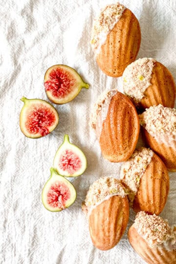 Golden brown spiced madeleines with a soft shell pattern and a partial dip into a white glaze and then a coating of freeze-dried figs all arranged on a white linen cloth.
